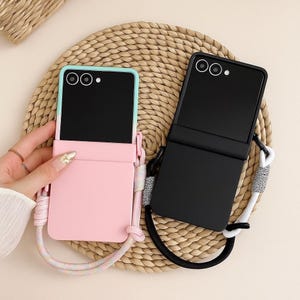 Puede incluir: Dos fundas para teléfono, una rosa y otra negra, con cordones adjuntos. La funda rosa tiene un borde azul claro y una bolsa rosa a juego. La funda negra tiene un cordón negro con detalles blancos. Ambas fundas están abiertas.