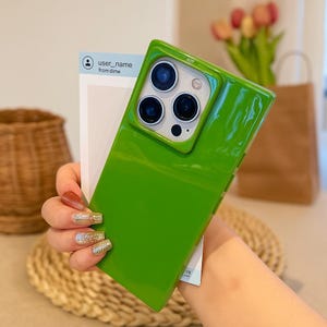 May include: A bright green phone case with a glossy finish. The case has a square camera cutout with three lenses. A white card with the text "user_name from dime" is held behind the phone. The phone case is held by a person.