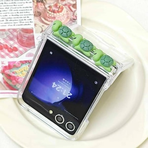 May include: Clear phone case with four green turtle embellishments. The phone screen displays the time as 21:24. The case is designed to fit a foldable phone, with a camera and flash visible.