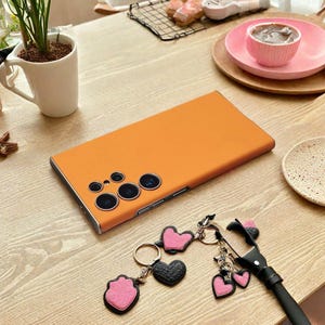 May include: An orange phone case with multiple camera lenses is displayed on a wooden surface. A collection of heart-shaped keychains in pink and black are next to the phone case. A cup of coffee and a small plant are in the background.