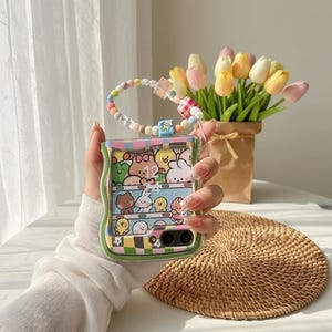 May include: A clear phone case with a colourful cartoon design featuring various animals and the number 12. The case has a wavy green border and a beaded wrist strap with pastel-coloured beads. A bouquet of tulips sits in the background.