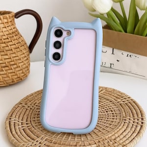 May include: A light blue phone case with cat ear accents. The case has a light pink interior and a camera module with three lenses. The phone case is sitting on a woven, circular placemat.