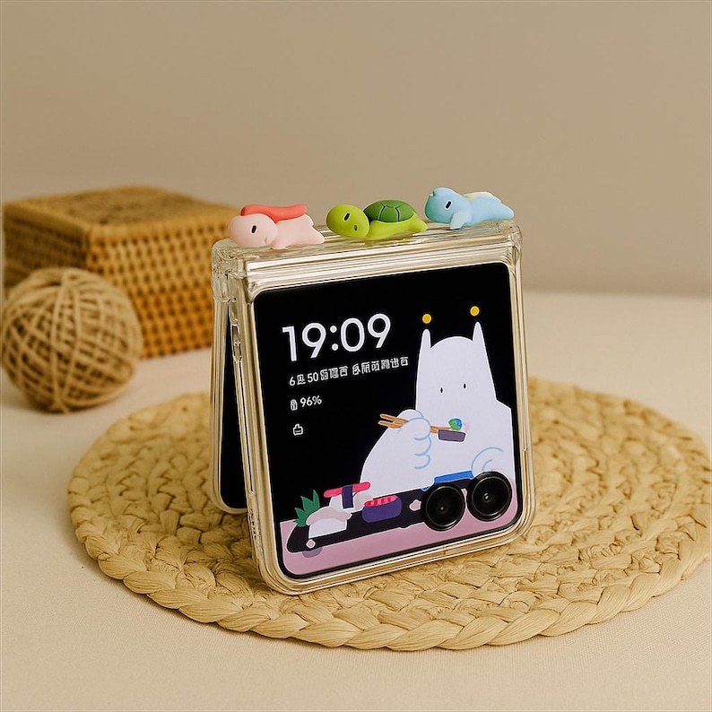 May include: A clear phone case with a screen displaying the time 19:09 and a cartoon character. The case is decorated with three small, colorful animal figurines: a pink creature, a green turtle, and a blue creature. The phone rests on a woven mat.