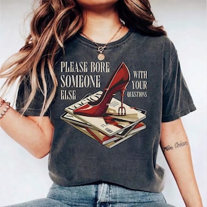 May include: Dark gray t-shirt with the text "PLEASE BORE SOMEONE ELSE WITH YOUR QUESTIONS." A red high heel shoe graphic is on top of a stack of magazines.
