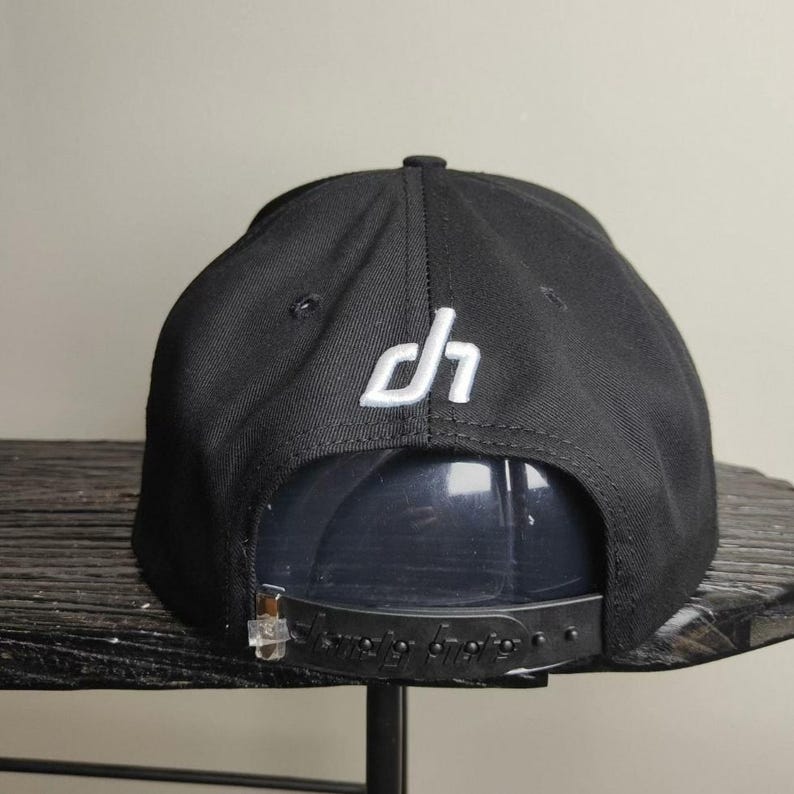 May include: Black snapback hat with a white embroidered "dh" logo on the back. The hat has a flat brim and a plastic snap closure for adjustable sizing. The hat is made of a durable fabric.