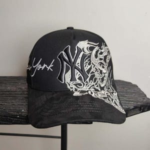 May include: Black baseball cap featuring the New York Yankees logo in white and a skull design. The cap has the word "York" embroidered in white script. The brim is a darker shade of black.