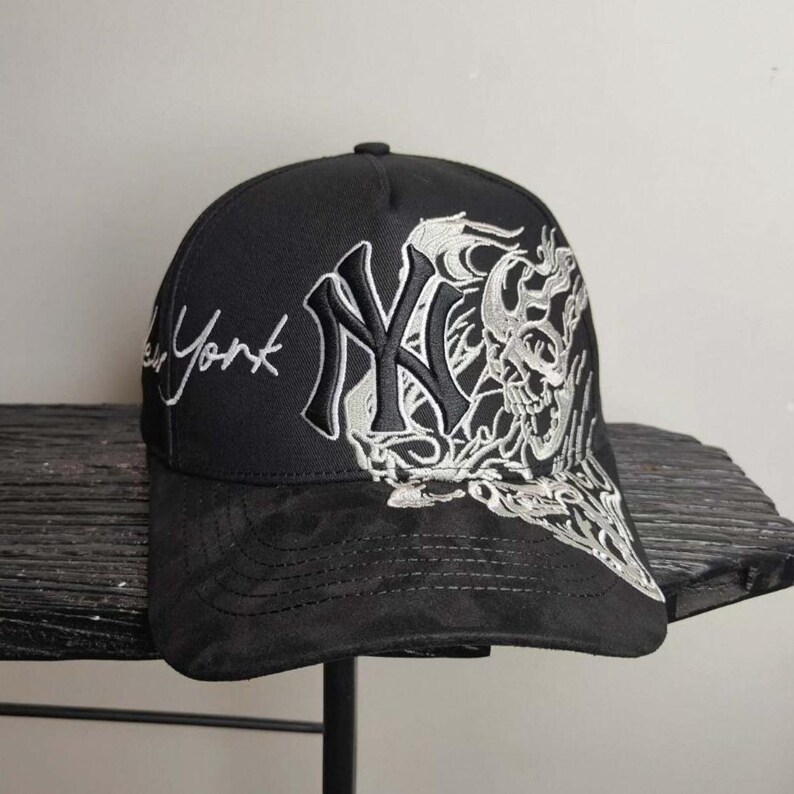 May include: Black baseball cap featuring the New York Yankees logo in white and a skull design. The cap has the word "York" embroidered in white script. The brim is a darker shade of black.