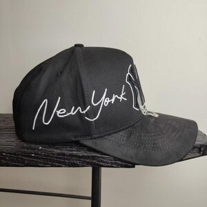 May include: Black baseball cap with the text "New York" in white script. The cap features the New York Yankees logo in white on the side. The hat has a curved brim and a structured crown.