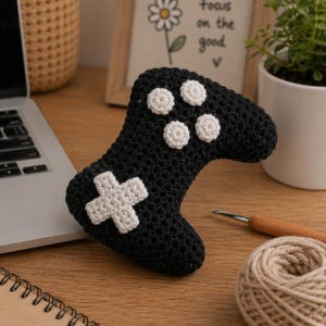 May include: A handmade black crochet video game controller with white buttons and a directional pad. The controller is placed on a wooden surface, with a laptop and a small potted plant in the background.