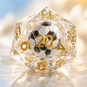 Könnte beinhalten: Ein klarer, vielseitiger Würfel mit einem Fußball im Inneren. Der Würfel hat goldfarbene Zahlen und ein Fußball-Design. Die Zahl 20 ist deutlich sichtbar. Dieser einzigartige Würfel ist ideal für Tischspiele.