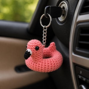 May include: A pink crocheted flamingo keychain with a black beak and eyes, and a white accent on the beak. The keychain is attached to a silver chain and ring, and is hanging near a car's ignition.