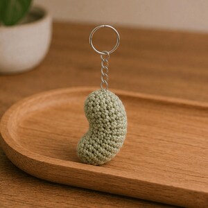 May include: A handmade, light green crochet bean-shaped keychain. The bean is attached to a silver-colored metal keyring and chain. The keychain is sitting on a wooden tray.