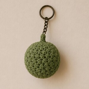 May include: A green crocheted keychain featuring a round, textured design. The keychain has a dark metal ring and chain. The crocheted sphere is a solid, muted green color.