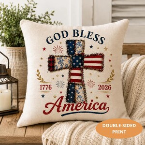 May include: A square, off-white pillow with a patriotic design. The pillow features a cross made of patches in red, white, and blue, with the words "GOD BLESS AMERICA" and the years "1776" and "2026". Decorative stars and fireworks complete the design.