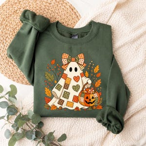 May include: An olive green crewneck sweatshirt featuring a graphic of a ghost wearing a patchwork quilt pattern with a matching bow. The ghost holds a pumpkin-shaped bucket filled with candy corn, surrounded by autumn leaves and berries.