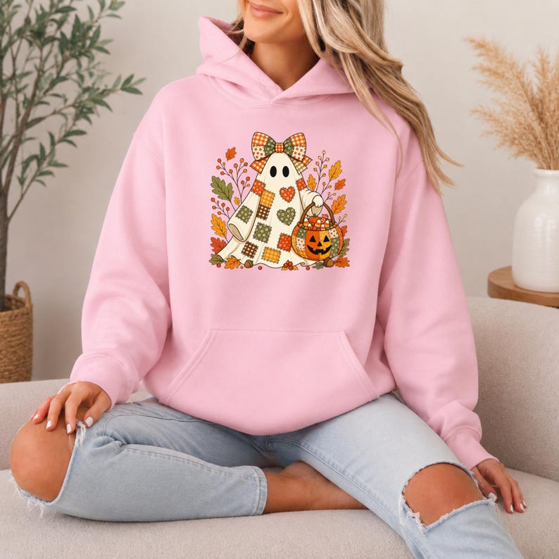 May include: A light pink hooded sweatshirt featuring a graphic of a ghost wearing a plaid bow, holding a jack-o'-lantern bucket filled with candy, surrounded by autumn leaves and hearts in shades of orange, green, and brown.