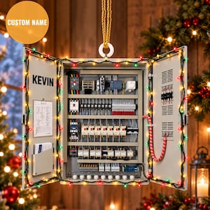 Puede incluir: Un adorno colgante con forma de cuadro eléctrico que lleva el nombre KEVIN impreso en la puerta interior. La caja metálica está decorada con luces de colores y contiene varios disyuntores, bloques de terminales y cableado interno.