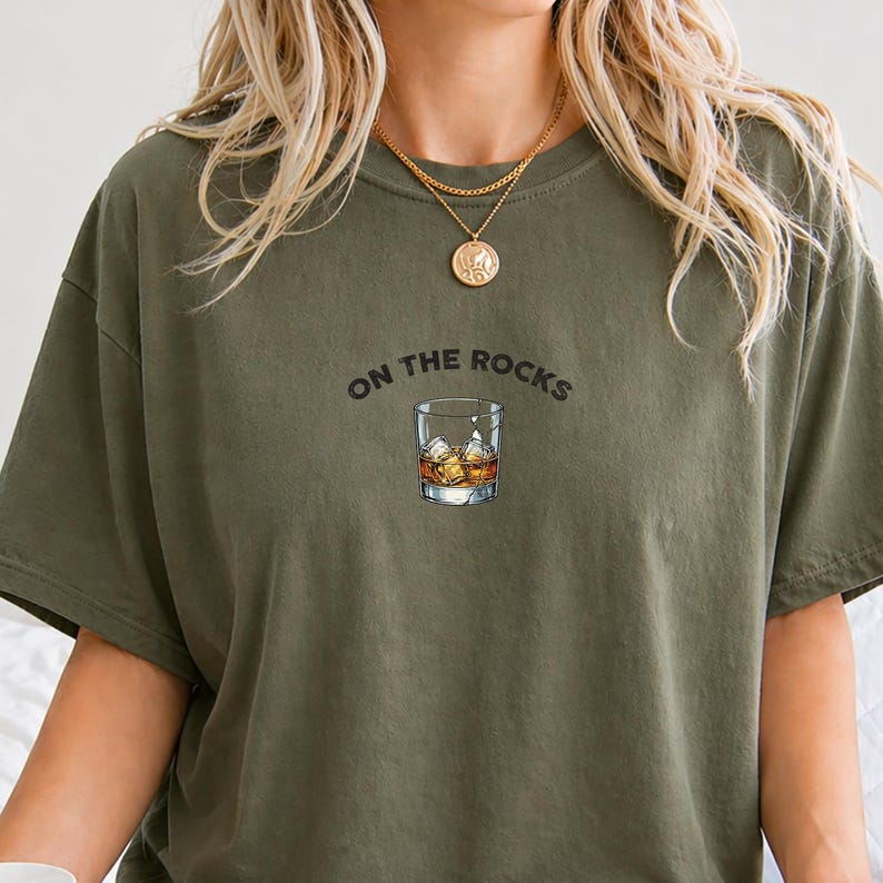 May include: Olive green t-shirt with the phrase "ON THE ROCKS" above a graphic of a whiskey glass filled with ice and amber liquid. The shirt has a crew neck and short sleeves.
