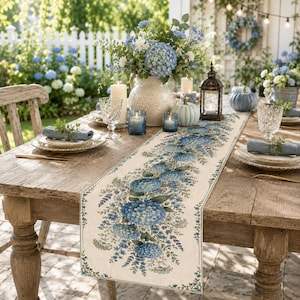 May include: A rectangular table runner featuring a printed pattern of blue hydrangea flowers and green foliage on a cream-colored fabric base. The runner is displayed on a wooden table with place settings, a floral centerpiece, and candles.