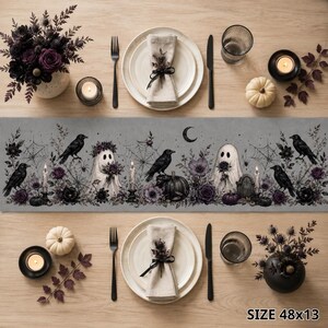 May include: A gray table runner featuring a repeating pattern of ghosts, black crows, spiderwebs, pumpkins, and dark purple flowers. The runner is displayed on a wooden table with place settings, including white plates, napkins, and candles. Size 48x13.