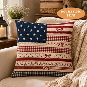 May include: A square throw pillow featuring a patriotic design with a blue section containing white stars and horizontal stripes in red, white, and blue. The pattern includes gingham, floral, and lace-like accents with small red bows. Text reads DOUBLE-SIDED PRINT.