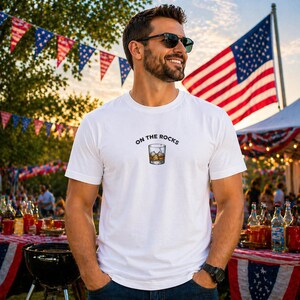 May include: White t-shirt with the text "ON THE ROCKS" above a graphic of a whiskey glass with ice. The background includes an American flag, festive bunting, and a table with drinks.