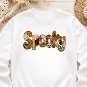 May include: A white crewneck sweatshirt featuring the word SPOOKY printed across the chest. Each letter is filled with a patchwork design of various autumn and Halloween-themed patterns, including plaid, leopard print, pumpkins, bats, and ghosts.