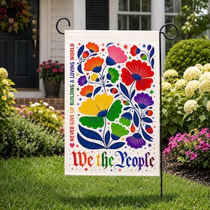 May include: A rectangular garden flag featuring a vibrant, multi-colored floral pattern with red, orange, yellow, green, blue, and purple petals. The text reads "NEVER GIVE UP BUILDING A LOVING WORLD" vertically and "We the People" in a stylized font.