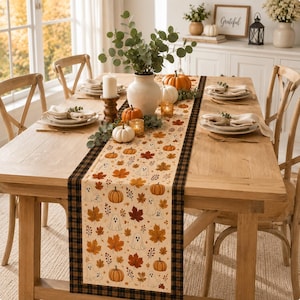 May include: A rectangular table runner featuring a cream-colored center with a repeating pattern of orange pumpkins, brown autumn leaves, and white ghosts, bordered by a black and brown plaid trim, displayed on a wooden dining table.