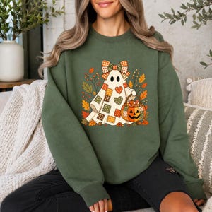 May include: An olive green crewneck sweatshirt featuring a graphic of a white ghost wearing a plaid bow and holding a pumpkin-shaped basket filled with candy corn, surrounded by colorful autumn leaves and small heart-shaped patches.