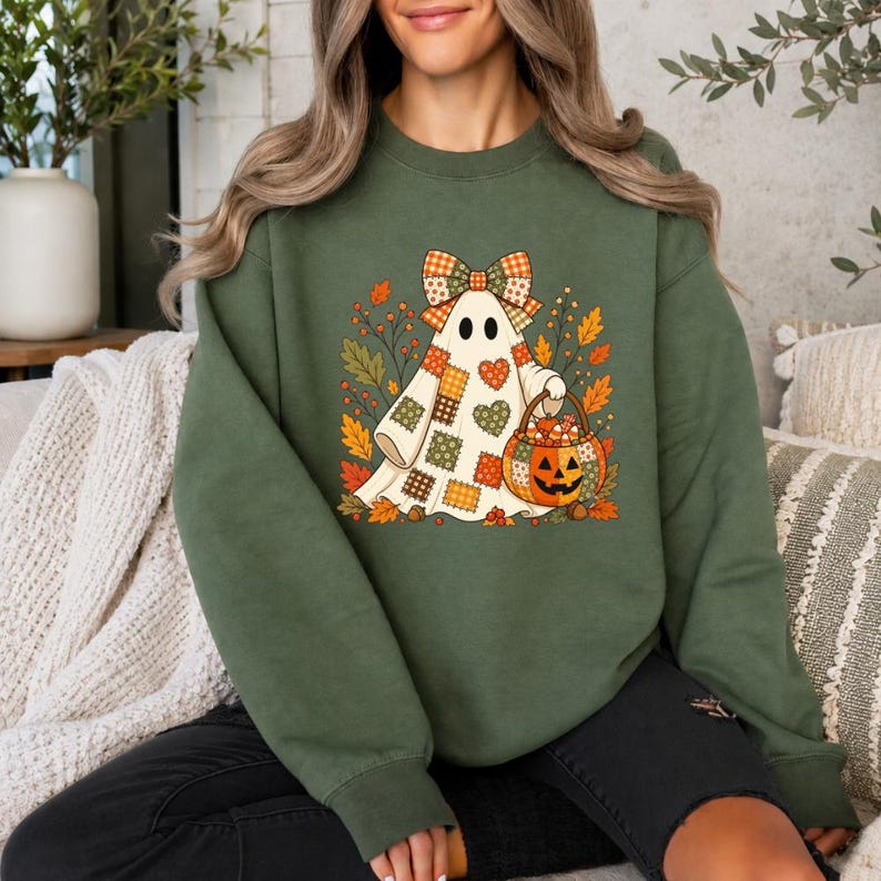 May include: An olive green crewneck sweatshirt featuring a graphic of a white ghost wearing a plaid bow and holding a pumpkin-shaped basket filled with candy corn, surrounded by colorful autumn leaves and small heart-shaped patches.