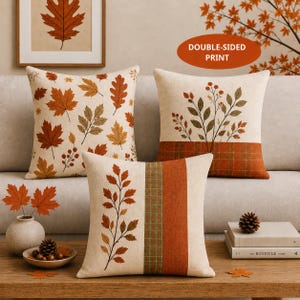 May include: Three square throw pillows featuring autumn-themed designs. One pillow has scattered orange and brown leaves, another displays a branch with berries above a plaid band, and the third shows a vertical leaf branch and stripes.