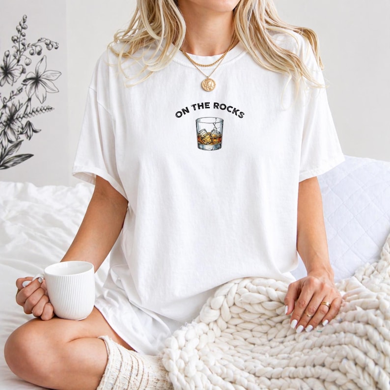 May include: White t-shirt with the phrase "ON THE ROCKS" above a graphic of a whiskey glass with ice cubes. The person is holding a white mug. The shirt is a casual, everyday wear item.