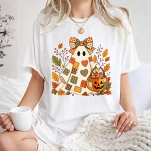 May include: A white short-sleeve t-shirt featuring a graphic of a ghost wearing a patchwork-style sheet with a large plaid bow on its head. The ghost holds a jack-o'-lantern bucket filled with candy, surrounded by autumn leaves and berries.