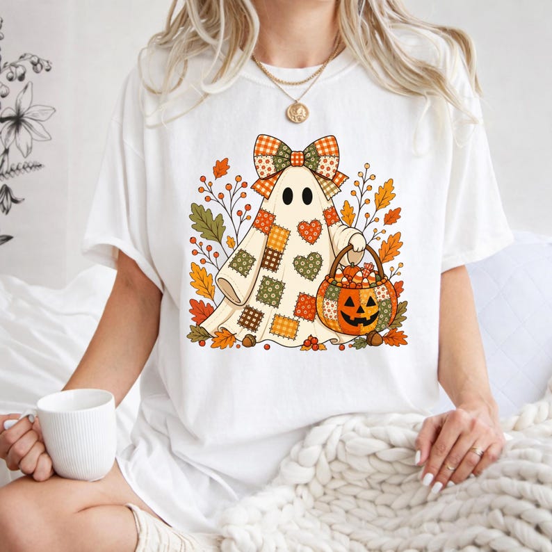 May include: A white short-sleeve t-shirt featuring a graphic of a ghost wearing a patchwork-style sheet with a large plaid bow on its head. The ghost holds a jack-o'-lantern bucket filled with candy, surrounded by autumn leaves and berries.