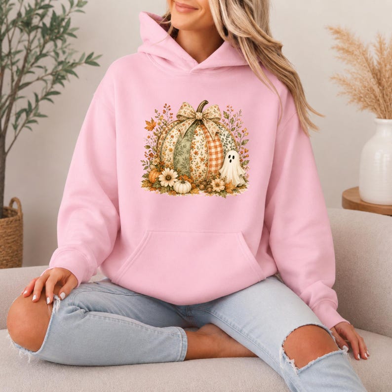 Peut inclure: Un sweat-shirt &agrave; capuche rose clair avec un motif central repr&eacute;sentant une grosse citrouille orn&eacute;e d'un n&oelig;ud &agrave; motifs, entour&eacute;e de feuilles d'automne, de petites fleurs et d'un fant&ocirc;me blanc. Le v&ecirc;tement poss&egrave;de des manches longues et une poche.