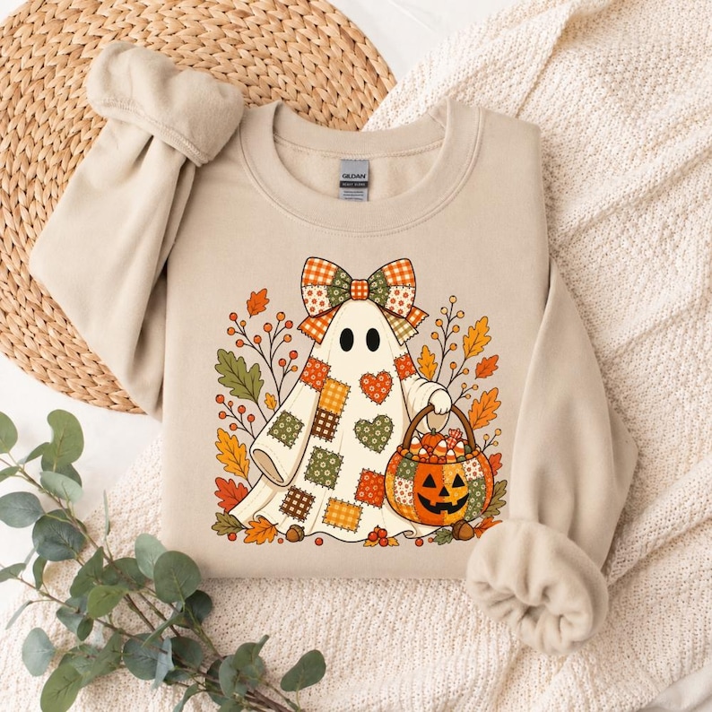 May include: A beige crewneck sweatshirt featuring a graphic of a ghost wearing a plaid bow and a patchwork-style sheet. The ghost holds a pumpkin-shaped basket filled with candy corn, surrounded by autumn leaves, berries, and acorns.