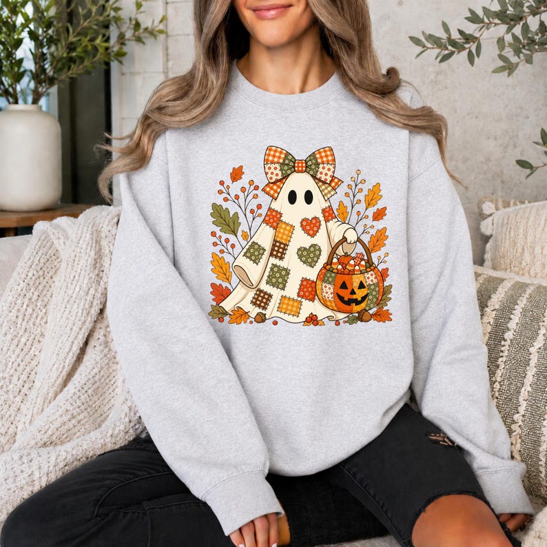 May include: A light gray crewneck sweatshirt featuring a graphic of a ghost wearing a plaid bow and a patchwork-style body. The ghost holds a jack-o'-lantern bucket filled with candy corn, surrounded by autumn leaves and branches.