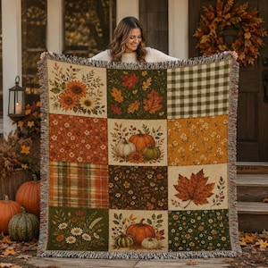 May include: A woven throw blanket featuring a nine-panel grid design with autumn-themed patterns including sunflowers, pumpkins, maple leaves, plaid, and gingham in shades of orange, green, brown, and cream with fringed edges on all four sides.