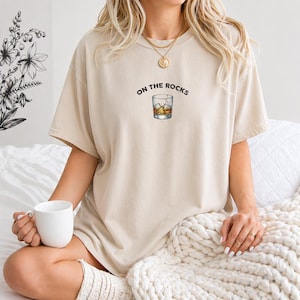 May include: Beige t-shirt with the phrase "ON THE ROCKS" above a graphic of a whiskey glass with ice cubes. The shirt is a neutral color and has a relaxed fit. The shirt is made of a soft material.