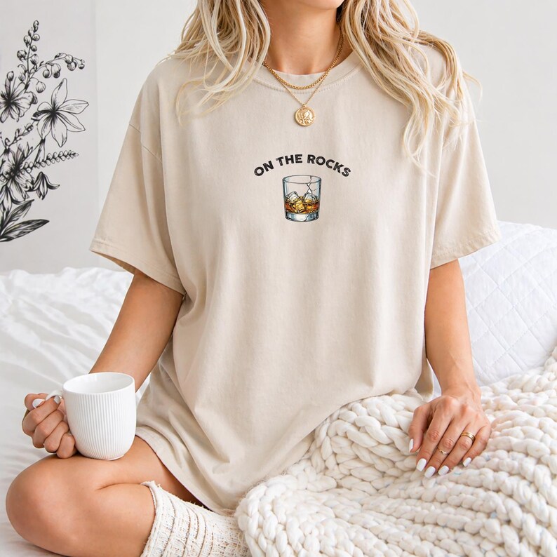 May include: Beige t-shirt with the phrase "ON THE ROCKS" above a graphic of a whiskey glass with ice cubes. The shirt is a neutral color and has a relaxed fit. The shirt is made of a soft material.