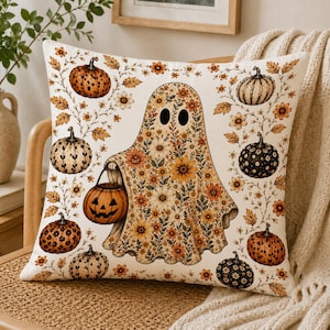 May include: A square throw pillow featuring a cream-colored background with a central ghost illustration covered in a floral pattern. The ghost holds a jack-o'-lantern bucket, surrounded by various pumpkins and orange and yellow flowers.