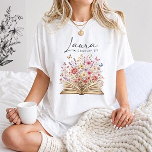 May include: A white short-sleeve t-shirt featuring a graphic of an open book with a bouquet of colorful wildflowers and butterflies emerging from the pages. The text "Laura" and "Chapter 57" is printed in black above the floral book illustration.