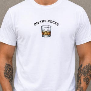 May include: White t-shirt with the phrase "ON THE ROCKS" above a graphic of a whiskey glass filled with ice and amber liquid. The design is centered on the chest.