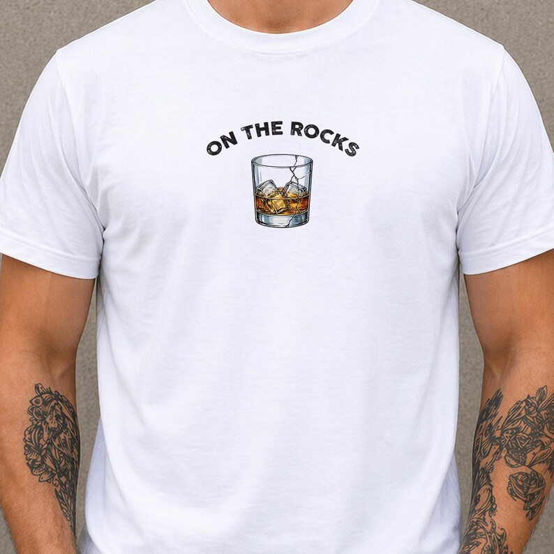May include: White t-shirt with the phrase "ON THE ROCKS" above a graphic of a whiskey glass filled with ice and amber liquid. The design is centered on the chest.