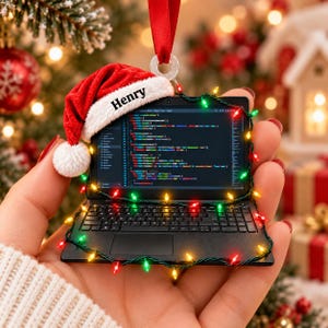 May include: A miniature black laptop ornament featuring a red Santa hat with the name Henry printed on the white trim. The screen displays lines of computer code, and the device is wrapped in a string of small, colourful Christmas lights.