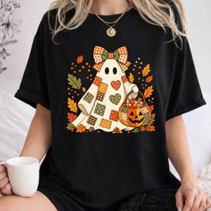 May include: A black t-shirt featuring a graphic of a ghost wearing a patchwork-style bow and holding a jack-o'-lantern bucket. The ghost is decorated with various heart and square patterns in orange, green, and brown, surrounded by autumn leaves.