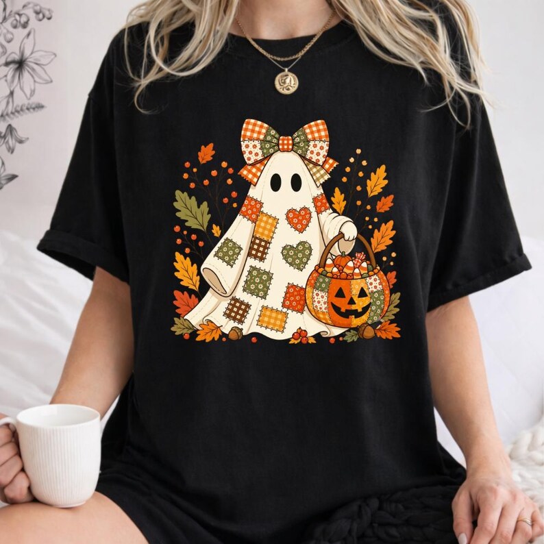 May include: A black t-shirt featuring a graphic of a ghost wearing a patchwork-style bow and holding a jack-o'-lantern bucket. The ghost is decorated with various heart and square patterns in orange, green, and brown, surrounded by autumn leaves.