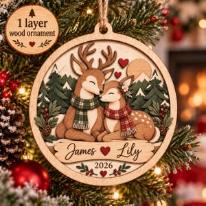 May include: A round wooden Christmas ornament featuring two deer wearing scarves, surrounded by pine trees and mountains. The ornament displays the names James and Lily with a heart in between, the year 2026, and a small circular tag reading 1 layer wood ornament.