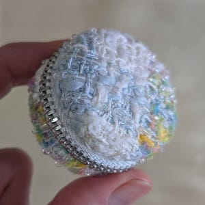 Op de afbeelding: Een klein, rond, met kralen bezet object met een rits. Het object is voornamelijk wit, lichtblauw en pastelkleuren. De rits is zilverkleurig. Het object wordt in de hand van een persoon gehouden.